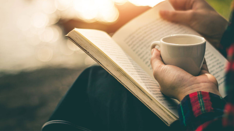 Persona che legge un libro tenendo una tazzina di caffè in mano 